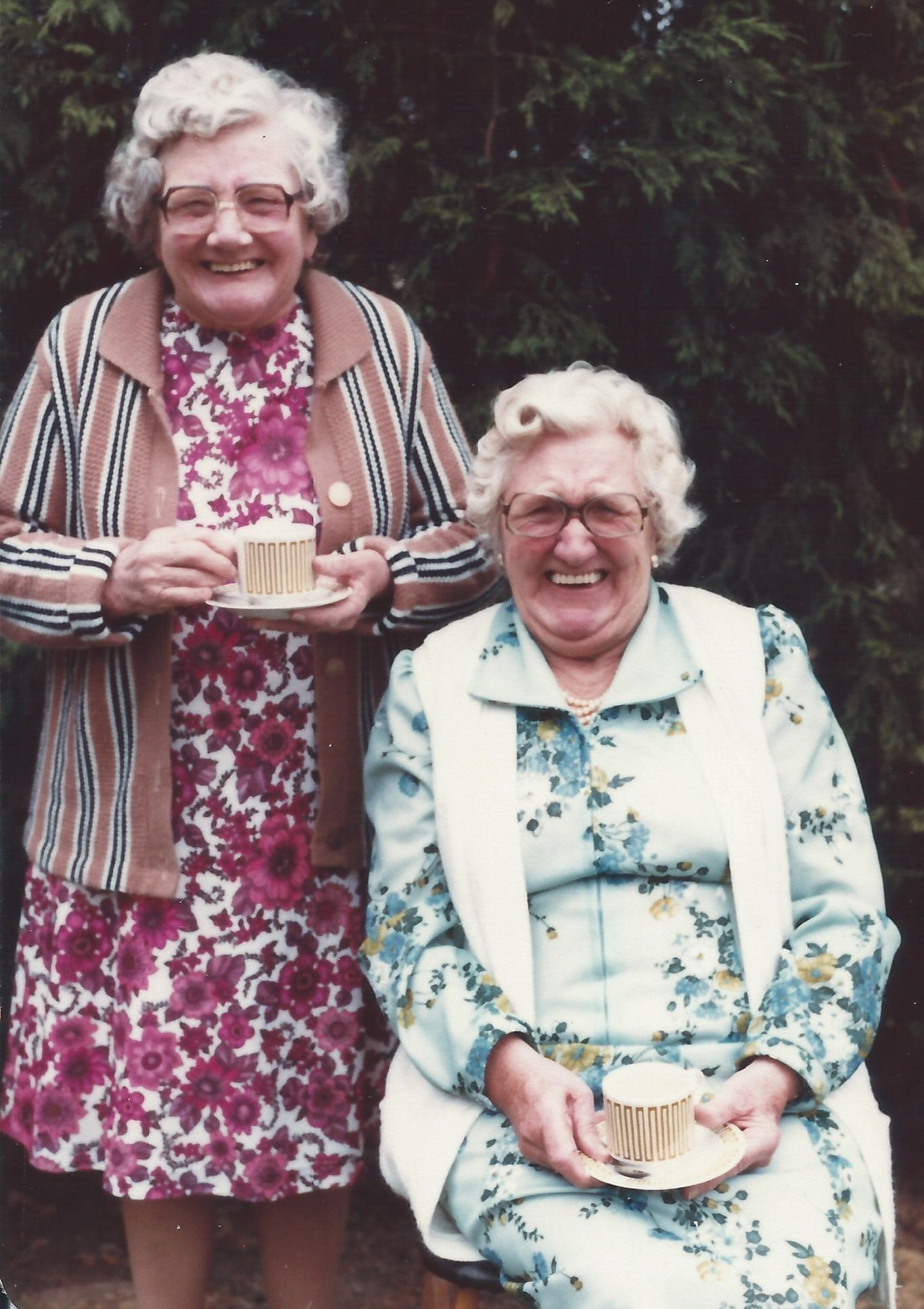 Two older ladies, one standing and one seated in front of a dark hedge. Both are smiling and holding a cup of tea