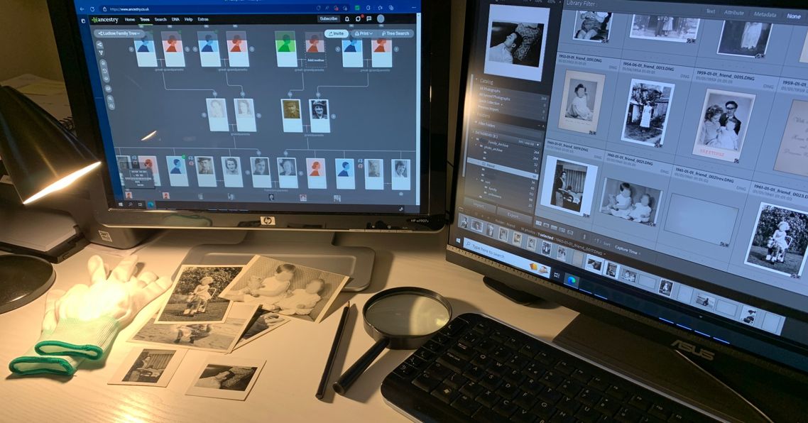 2 monitor screens display a family tree website & Adobe Lightroom. Both are on a desk with old photos, gloves, pencil and magnifying glass